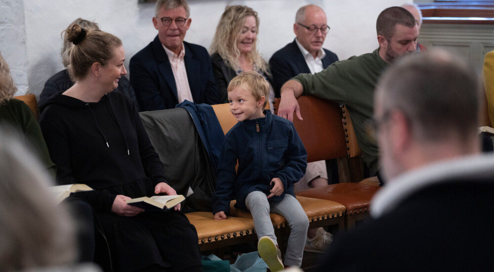 Lille dreng sidder på stole blandt voksne i kirke